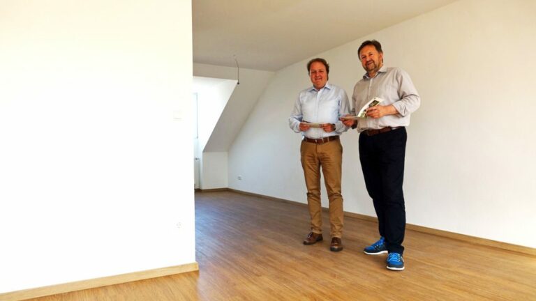 Zwei Männer stehen in einem leeren Raum mit Holzboden und halten Unterlagen in der Hand.
